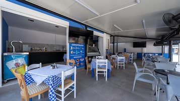 A restaurant interior with checkered tablecloths, white chairs, a blue and white color scheme, a kitchen area, and a sign advertising cocktails and desserts.