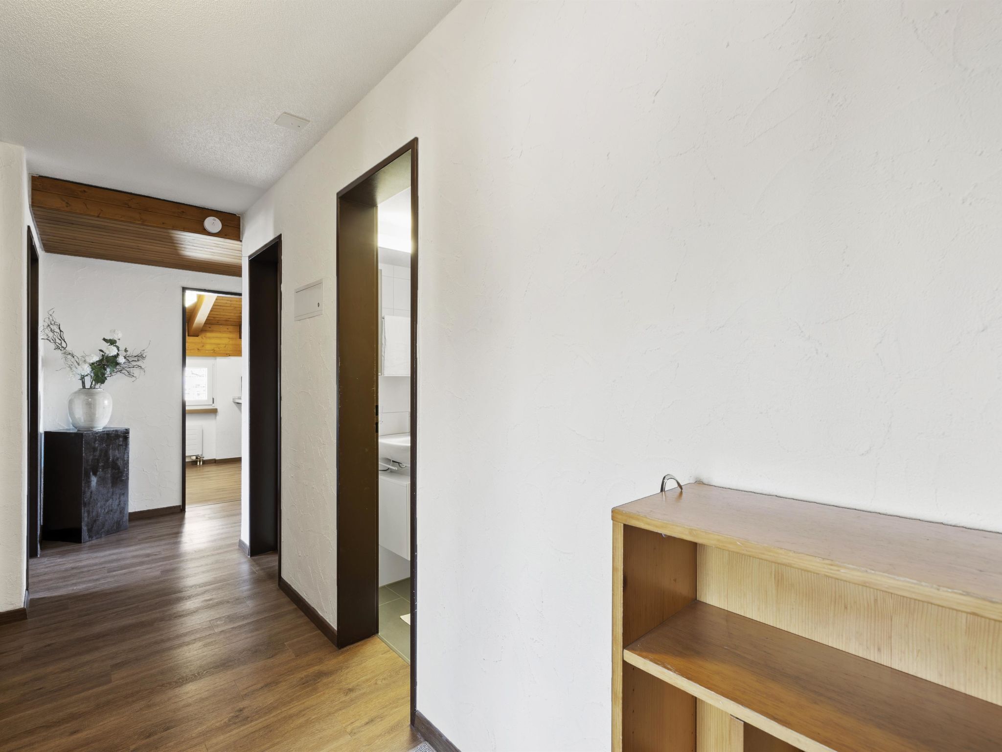 Een gang met houten vloer, een witte muur en een deur naar een andere kamer.