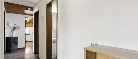 A hallway with wooden flooring, a white wall, and a door leading to another room.