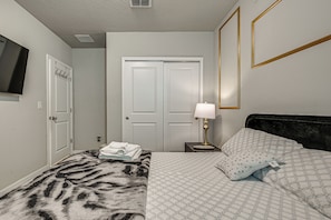 A bedroom with a bed, a bedside table with a lamp, a wall-mounted TV, and a door with a coat rack. - At Home Away From Home in Zhills (Zephyrhills)