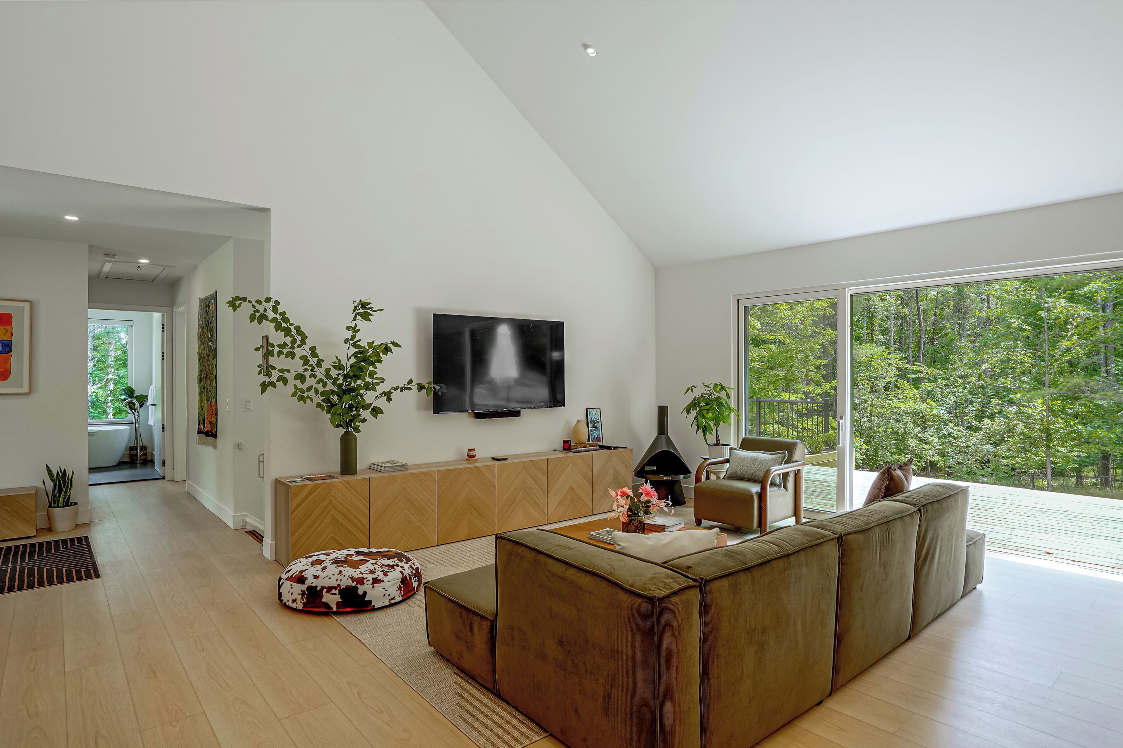 A modern living room with a large TV, wooden furniture, and a view of a forest through large windows.