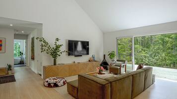 A modern living room with a large TV, wooden furniture, and a view of a forest through large windows.