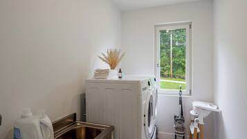 A laundry room with a washing machine, a sink, and a window with a view of greenery.