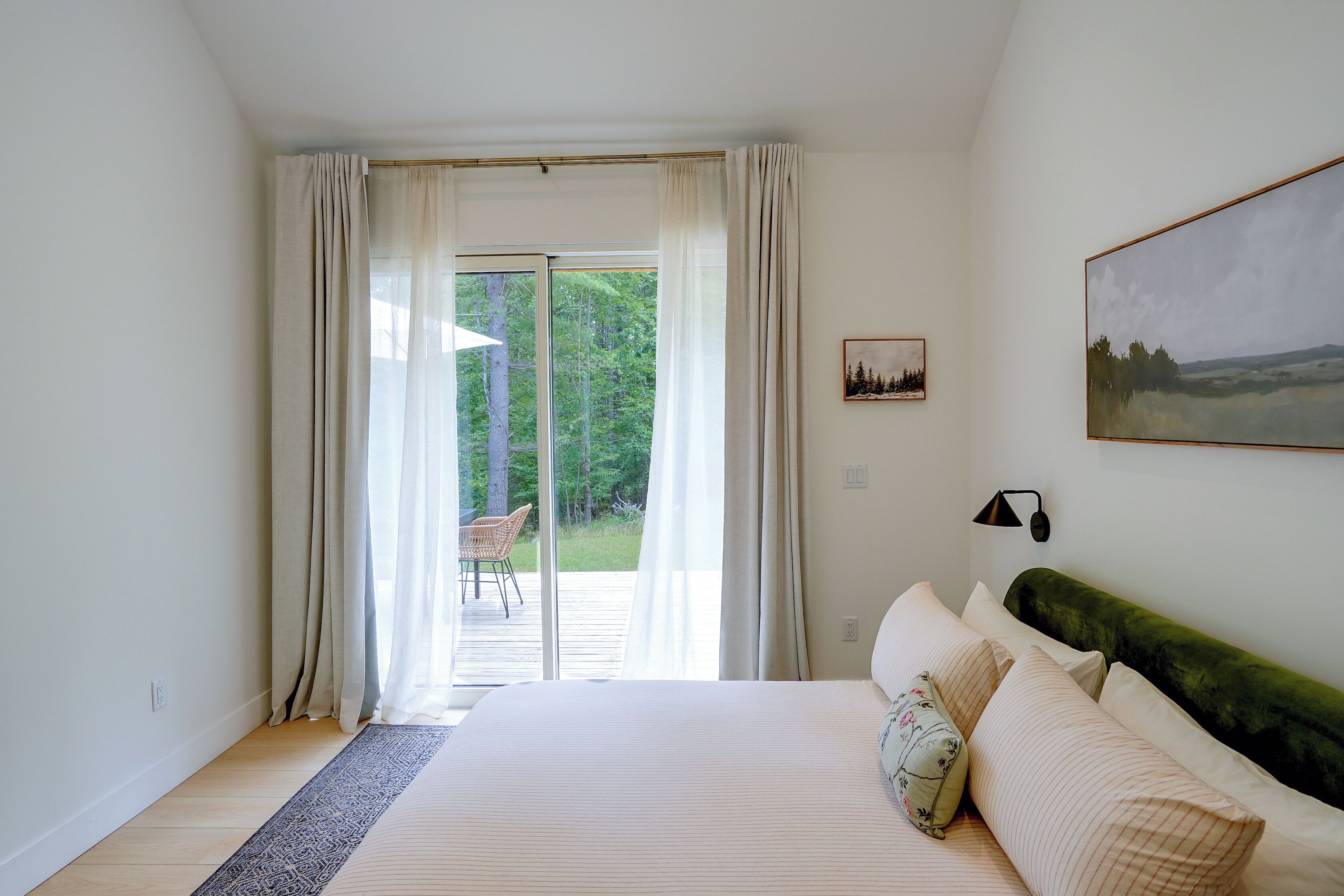 A bedroom with a large bed, a view of a forest through a sliding glass door, and a painting on the wall.
