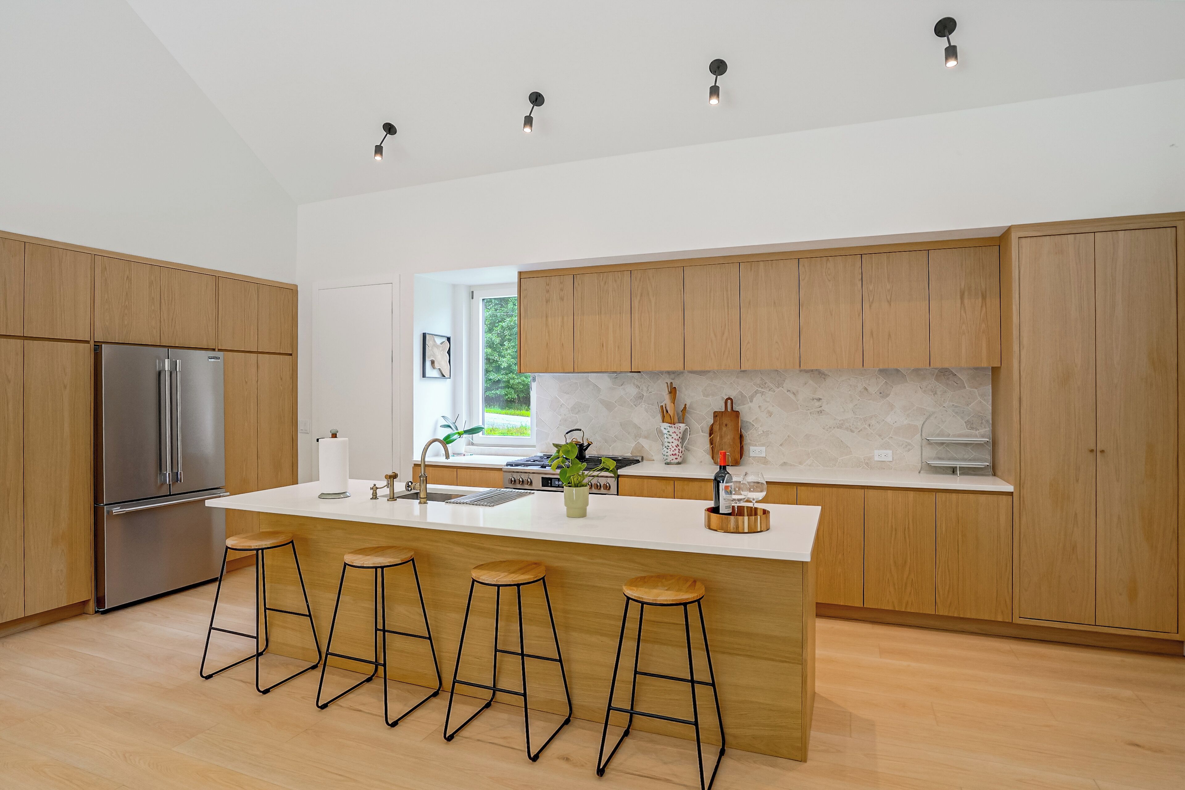 A modern kitchen with wooden cabinets, a stainless steel refrigerator, a large island with stools, and pendant lighting.