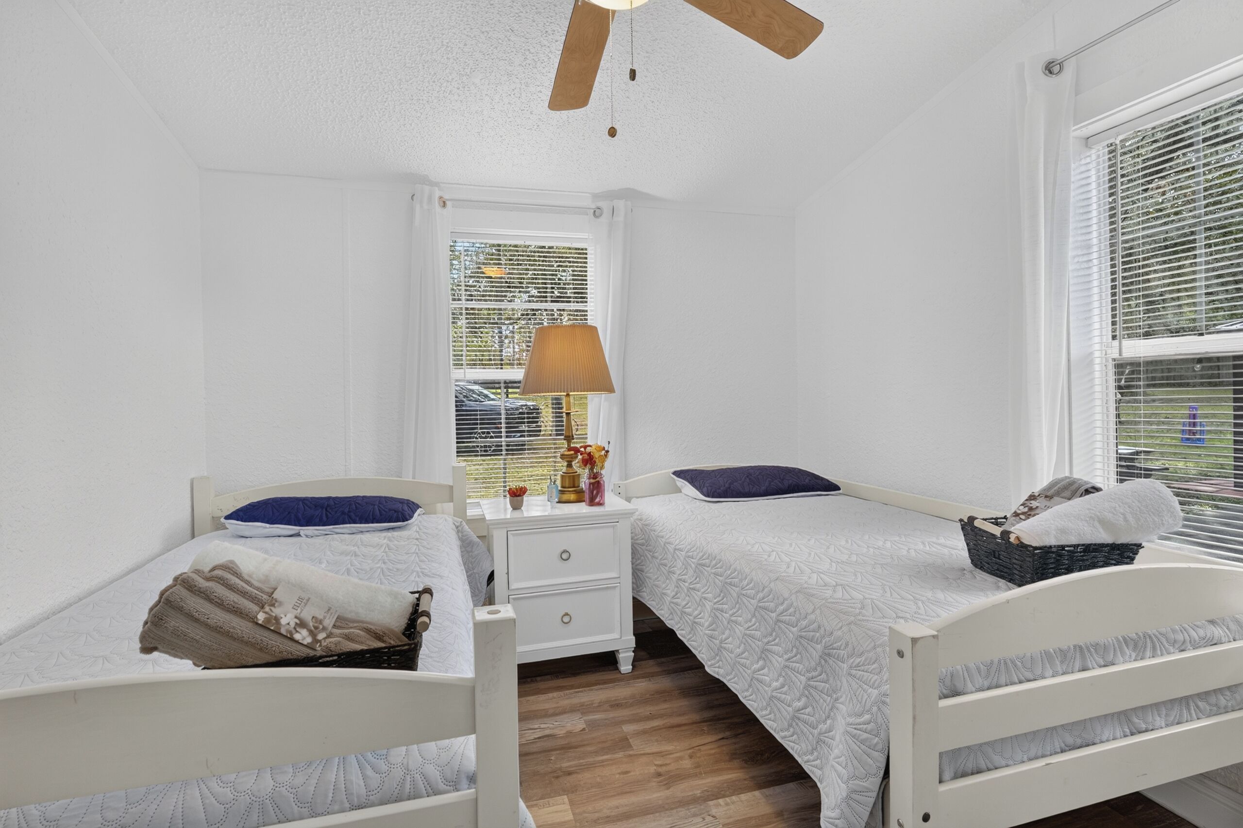 Une chambre avec deux lits simples, une table de chevet, une lampe, une fenêtre avec des stores et un ventilateur de plafond.