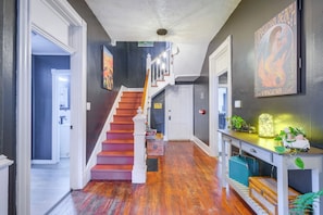 A hallway with a wooden staircase, a desk with plants, and a painting on the wall.
