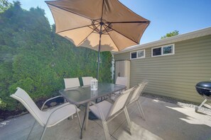 An outdoor patio with a glass table, white chairs, a large beige umbrella, and a barbecue grill. - Dual Cottage Escape Walkable to Gotl Strip (Geneva)