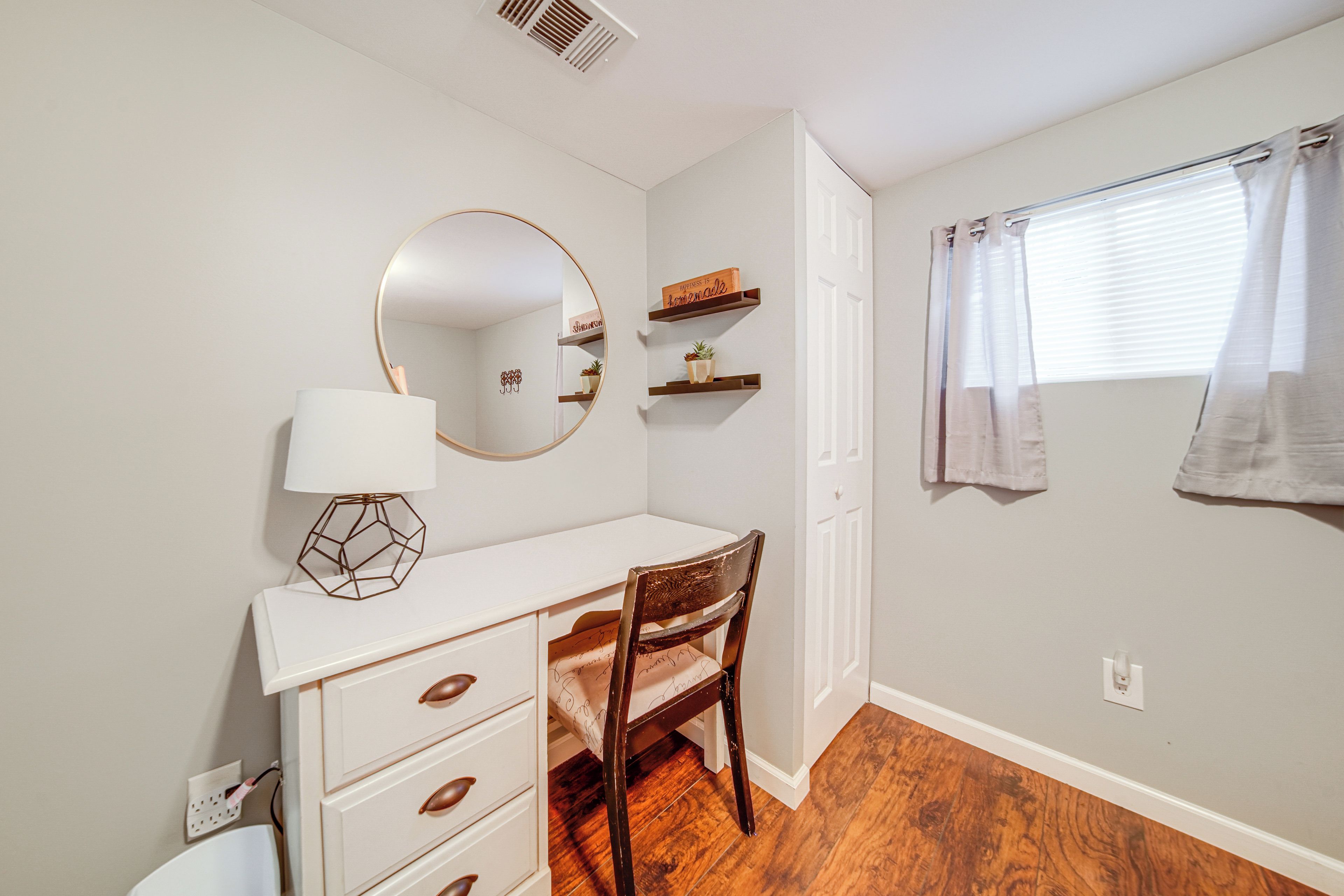 Une petite chambre à coucher, bien rangée, avec une commode blanche, un miroir rond, une chaise en bois et une fenêtre avec un rideau.