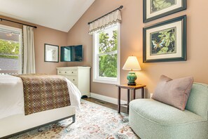 Ein Schlafzimmer mit Bett, Nachttisch, Lampe, Stuhl und gerahmten Bildern an der Wand.