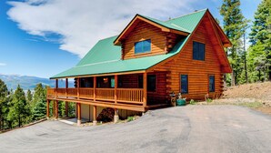 A two-story log cabin with a green roof, a porch, and a view of mountains.