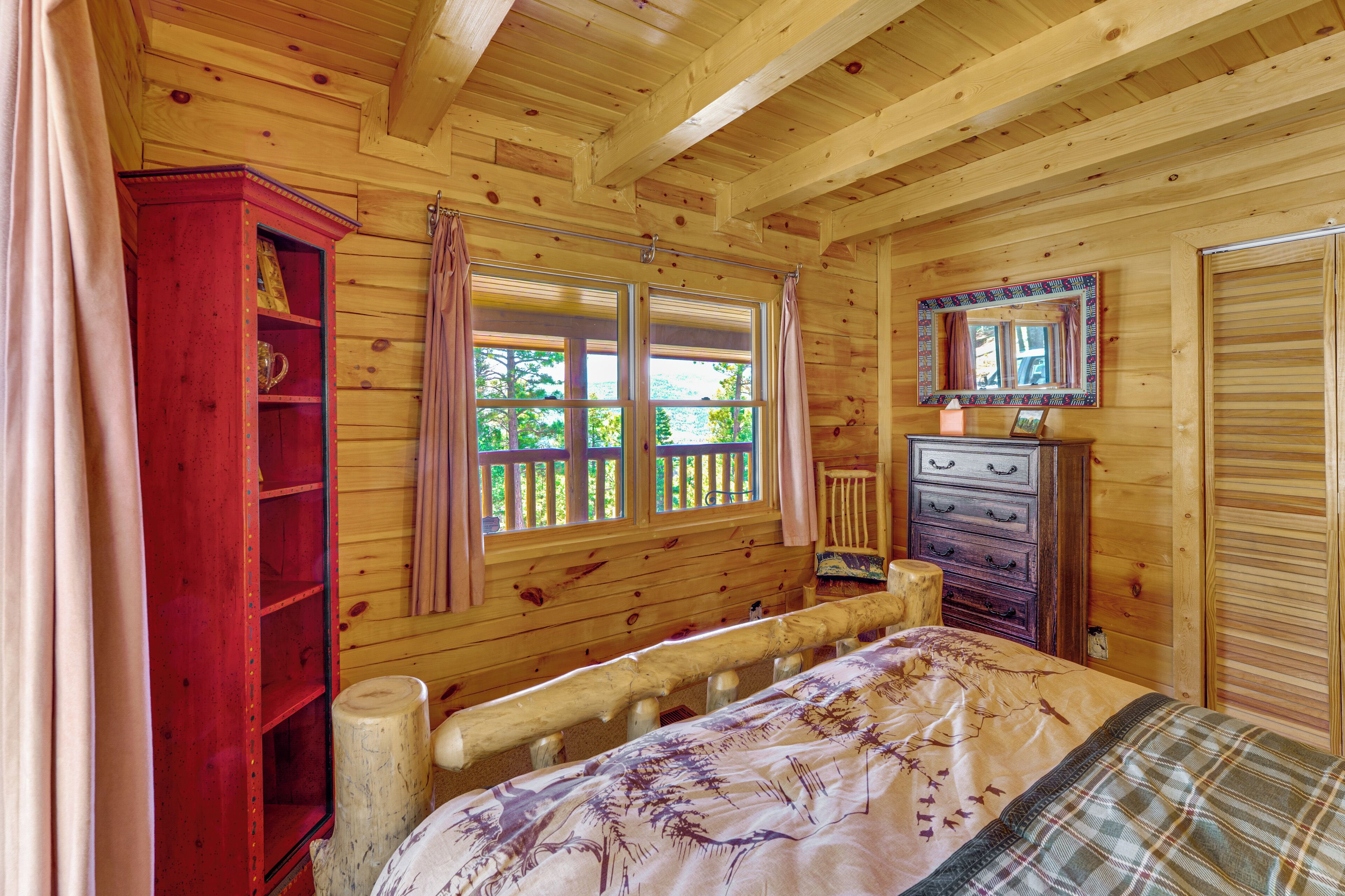 A wooden cabin room with a bed, a dresser, a mirror, and a window with curtains.