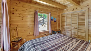 A wooden cabin bedroom with a bed, a window with curtains, a small table, and a wardrobe.