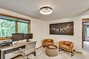 Una sala moderna con un escritorio, dos sillas, un pouf redondo y una gran obra de arte con una rama de árbol en la pared.