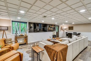 Ein großzügiger Wohnbereich mit einer großen, L-förmigen Couch, einem Couchtisch und einem Kamin. Es gibt einen Essbereich mit Tisch und Stühlen sowie einen an der Wand befestigten Fernseher.