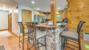 A kitchen with a stone island, wooden cabinets, and bar stools. - River Access! Wooded Cabin in Great Cacapon (Great Cacapon)