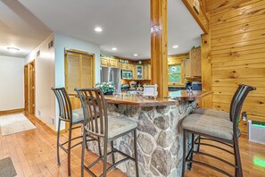 A kitchen with a stone island, wooden cabinets, and bar stools. - River Access! Wooded Cabin in Great Cacapon (Great Cacapon)