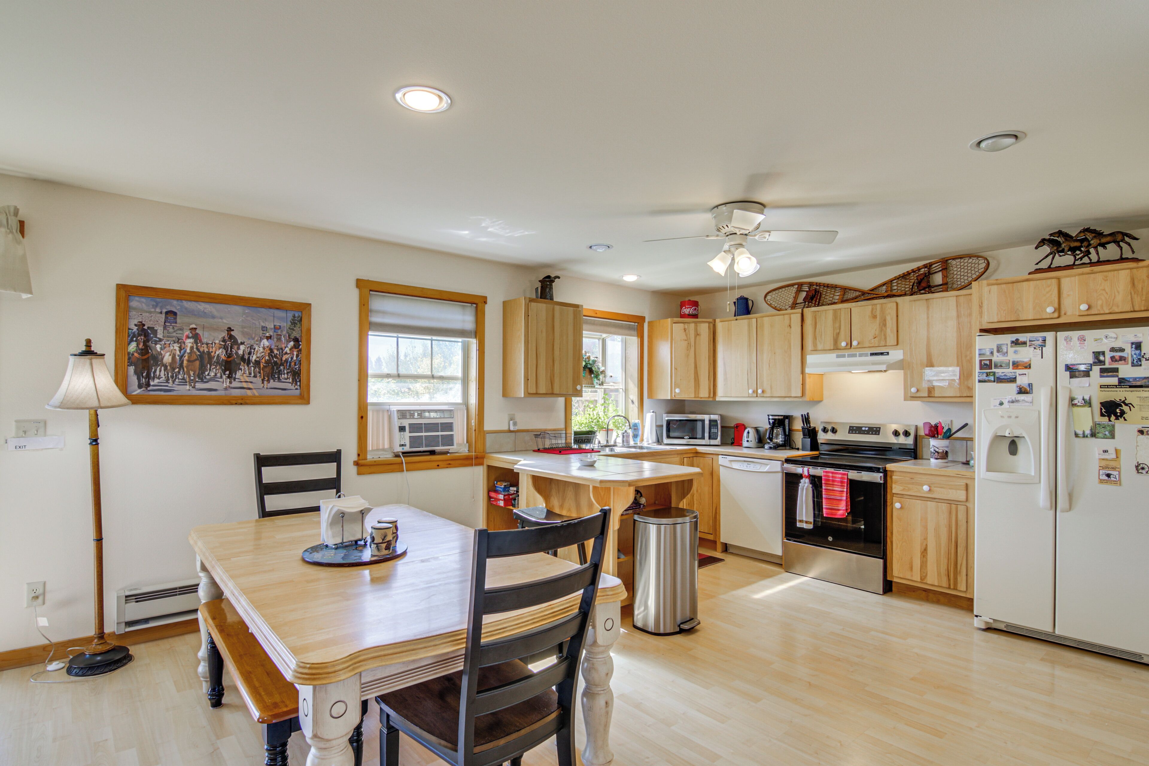 A kitchen with wooden cabinets, a dining table with chairs, a ceiling fan, and a refrigerator with magnets.