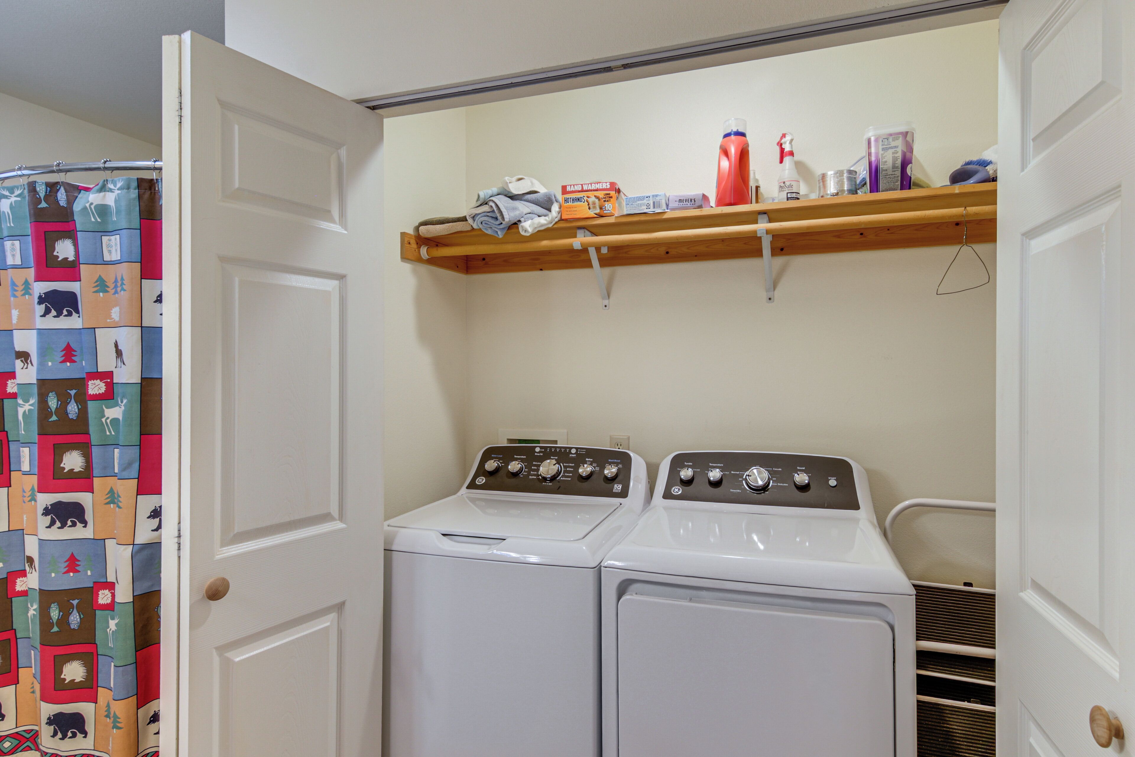 A laundry room with two white front-loading washing machines, a wooden shelf with various items, and a door with a colorful animal-themed curtain.
