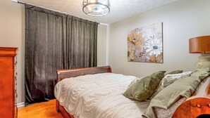 A bedroom with a wooden bed, a headboard, a nightstand, a lamp, a painting on the wall, and a window with curtains.