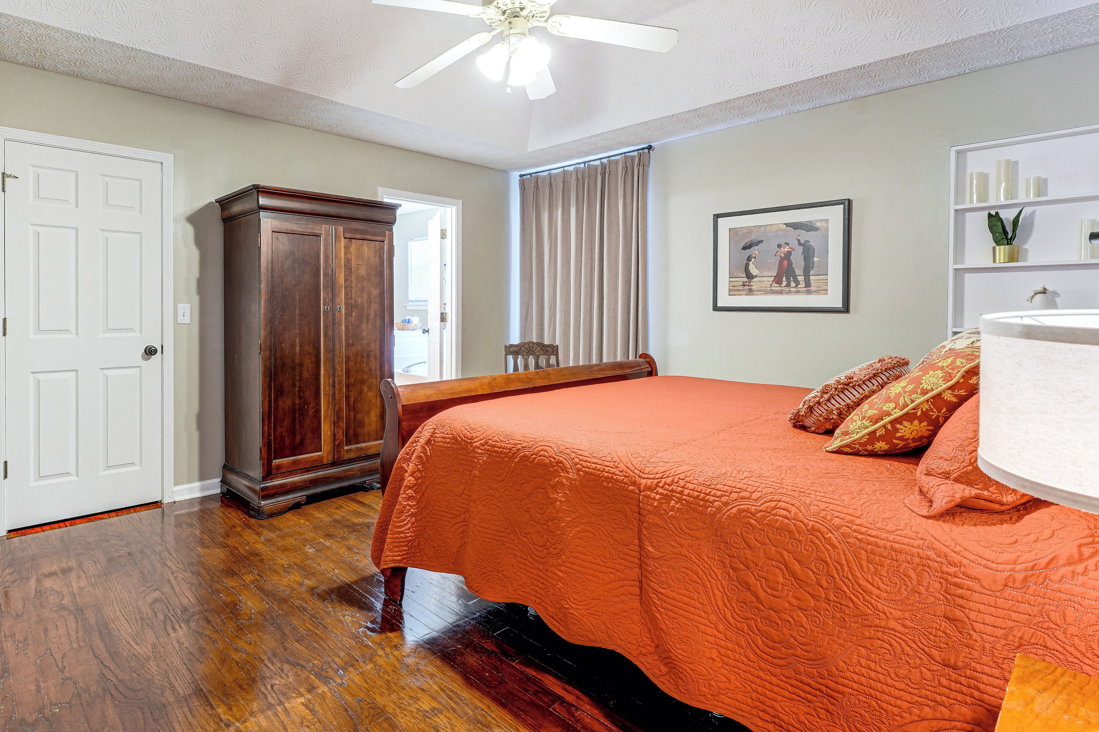 A bedroom with a wooden wardrobe, a bed with an orange bedspread, a ceiling fan, and a framed picture on the wall.