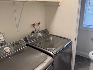 Ein Waschraum mit zwei übereinander gestapelten Waschmaschinen und Trocknern, einem Warmwasserbereiter und einem Fenster mit Jalousien.
