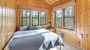 A wooden cabin bedroom with a bed, two windows, a bedside table with a lamp, and a heater. - Fish & Hike On-site! 4-cabin Pigeon River Retreat (Vanderbilt)