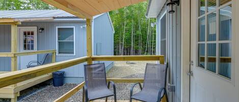 A wooden deck with two chairs, a small table, and a covered area with a window overlooking a forest.