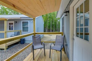 A wooden deck with two chairs, a small table, and a covered area with a window overlooking a forest.