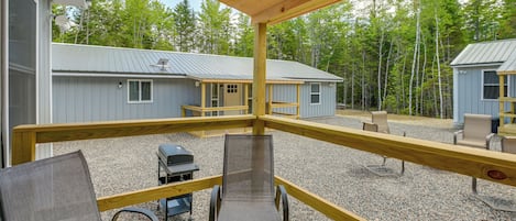 A covered outdoor deck with a grill and seating area, overlooking cabins and a forested area.