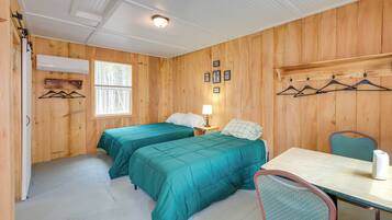 A room with two beds, a table, a chair, a window, and a wall-mounted shelf with pictures.