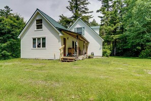 Ein weißes, zweistöckiges Haus mit Veranda, umgeben von einem grasbewachsenen Garten und Bäumen.
