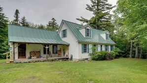 A two-story house with a green metal roof, a porch with outdoor furniture, and a well-maintained lawn. - Deck & Wooded Views! Rural Windham Group Retreat (West Townshend)