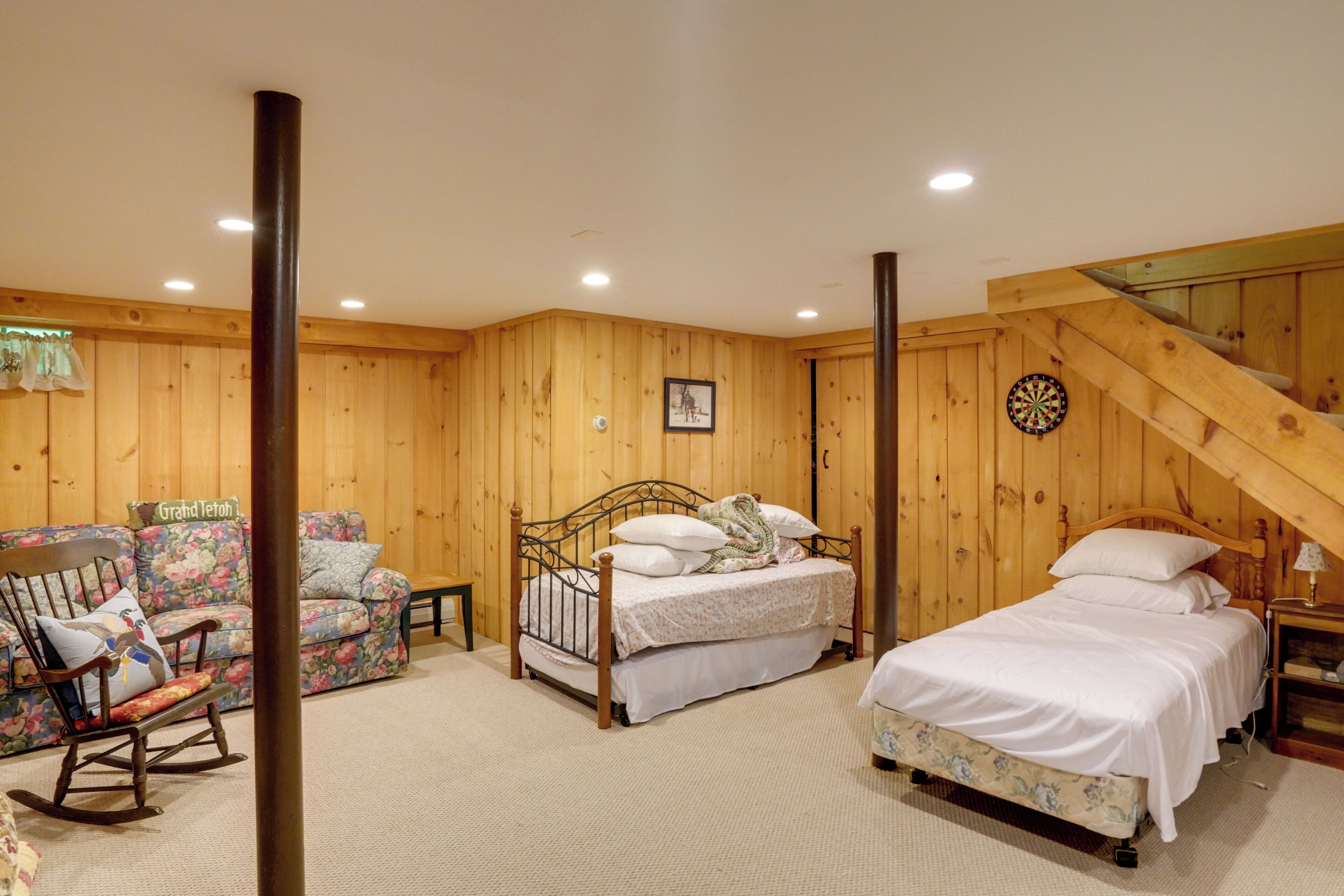 A room with wooden walls, a bed with white bedding, a rocking chair, and a dartboard.