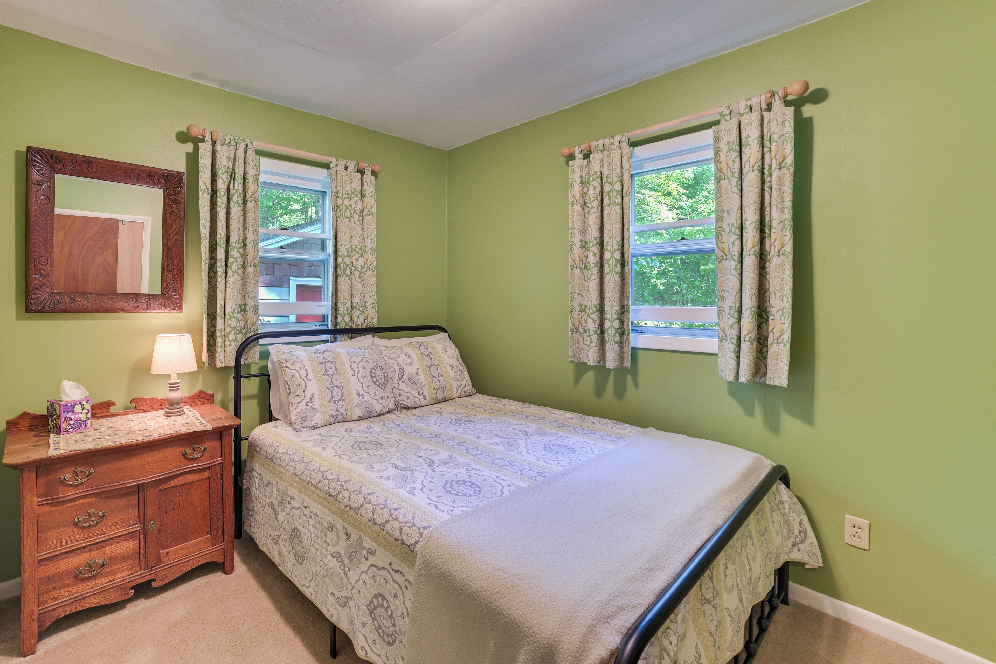 A bedroom with a bed, a nightstand, a mirror, and two windows with curtains.