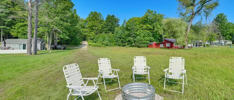 A grassy outdoor area with four white metal chairs arranged in a circle around a metal fire pit.