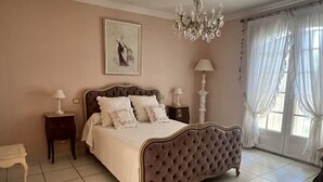 Een slaapkamer met een groot bed, twee nachtkastjes, een kroonluchter, een raam met gordijnen en een schilderij aan de muur.