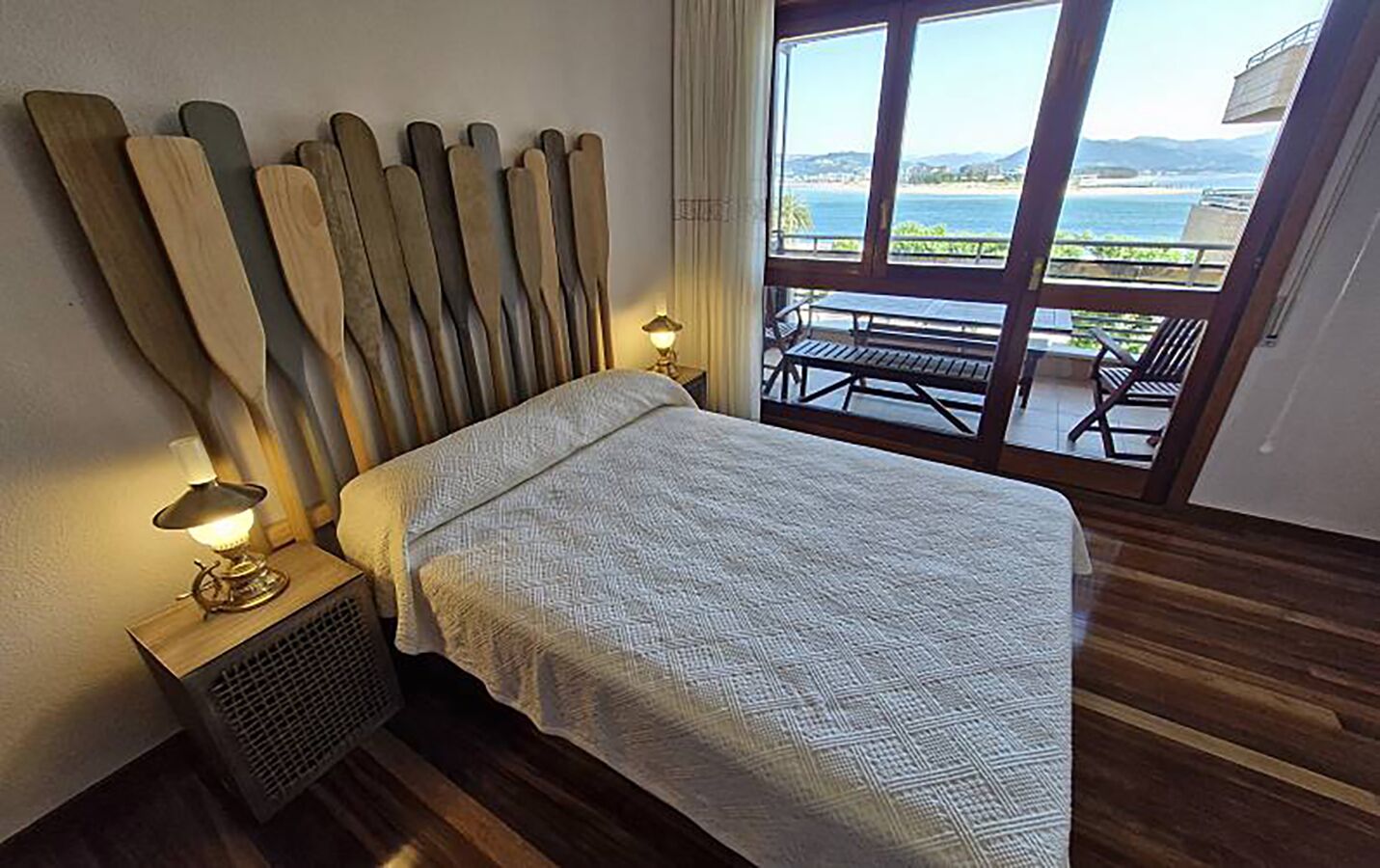 Un dormitorio con cama, una mesita de noche con lámpara, ventana con vista al mar y suelo de madera.