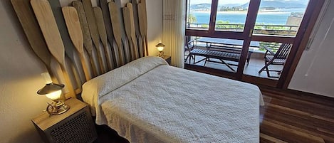 Un dormitorio con cama, una mesita de noche con lámpara, ventana con vista al mar y suelo de madera.