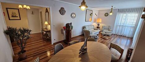 Une salle à manger avec une table ronde, des chaises et un lustre. On aperçoit un salon avec un canapé et une lampe au fond.
