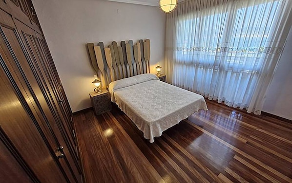 Un dormitorio con piso de madera, una cama con cubierta blanca, dos lámparas de noche y una ventana con cortinas translúcidas.