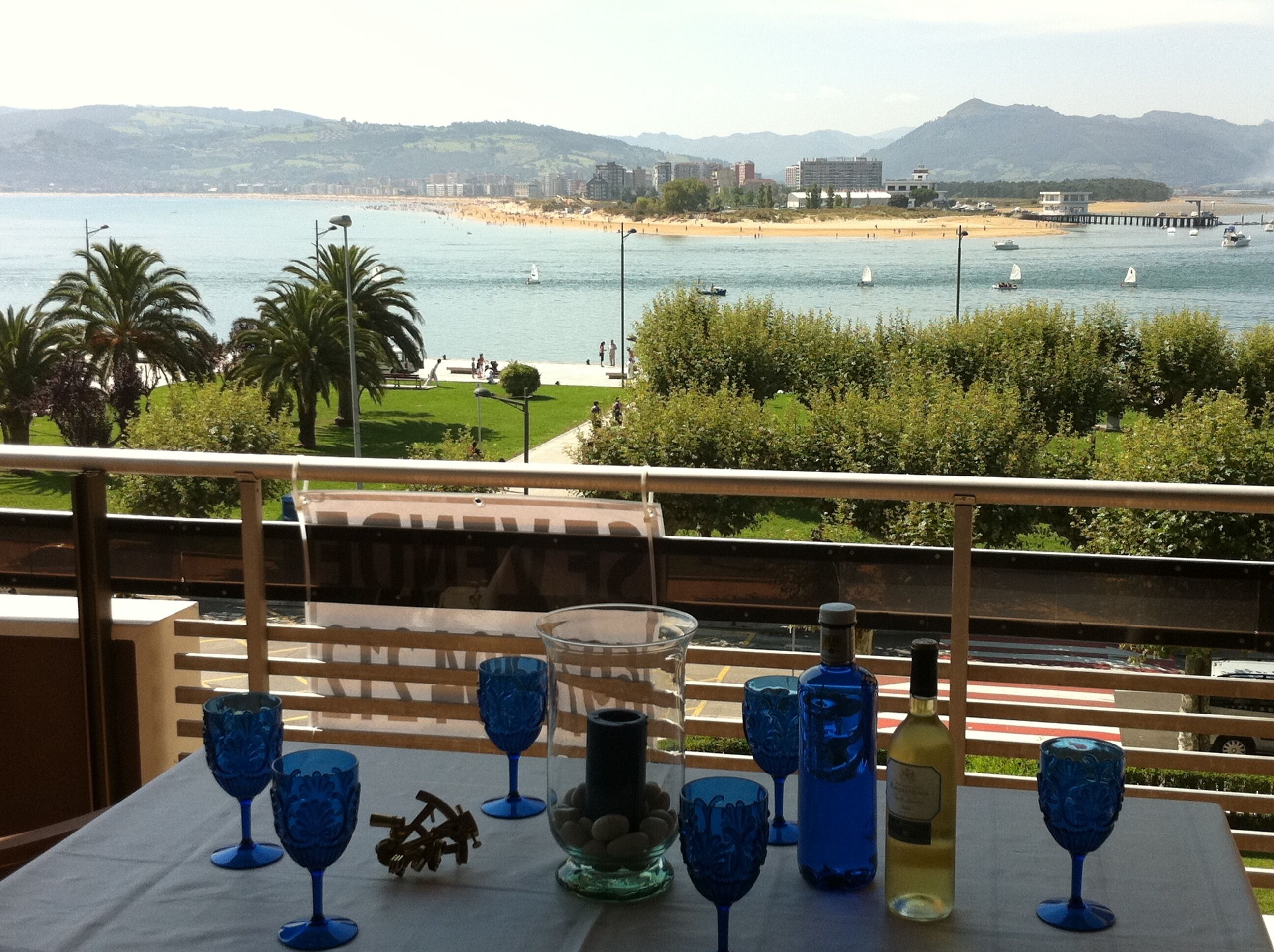 Mesa con cristalería azul y una botella, con vista a una playa y agua con barcos.
