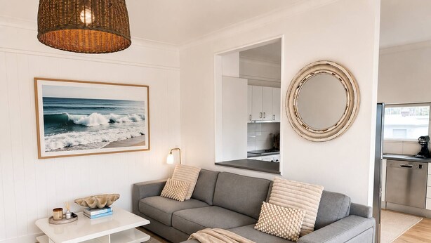 A modern living room with a grey sofa, a coffee table, a large framed picture of the ocean, a round mirror, and a kitchen in the background. - Coastal Retreat, 2 Bedroom Apartment Near Tathra Beach (Tathra)