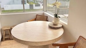 A round table with a woven base, surrounded by wooden chairs with woven backs, set in a room with large windows offering a view of a white building and greenery outside. - Coastal Retreat, 2 Bedroom Apartment Near Tathra Beach (Tathra)