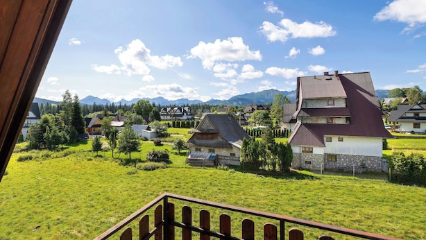 Ländliche Landschaft mit traditionellen Häusern, einem Holzzaun und einem Gebirgszug im Hintergrund.