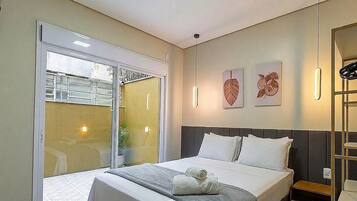 Quarto de hotel com uma cama, mesa de cabeceira, luminária na parede e uma porta de vidro deslizante para uma área externa.