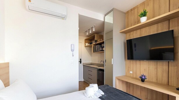 Een hotelkamer met een bed, een aan de muur bevestigde televisie, een klein keukentje en een houten plank met een plant.