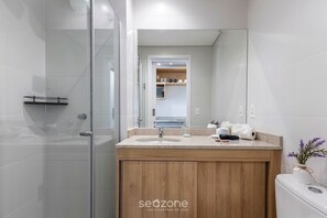 A modern bathroom with a glass shower enclosure, a wooden vanity with a sink, and a mirror reflecting a kitchen area. - Sophisticated Studio in a Cond. With a gym Man1707 (Curitiba)