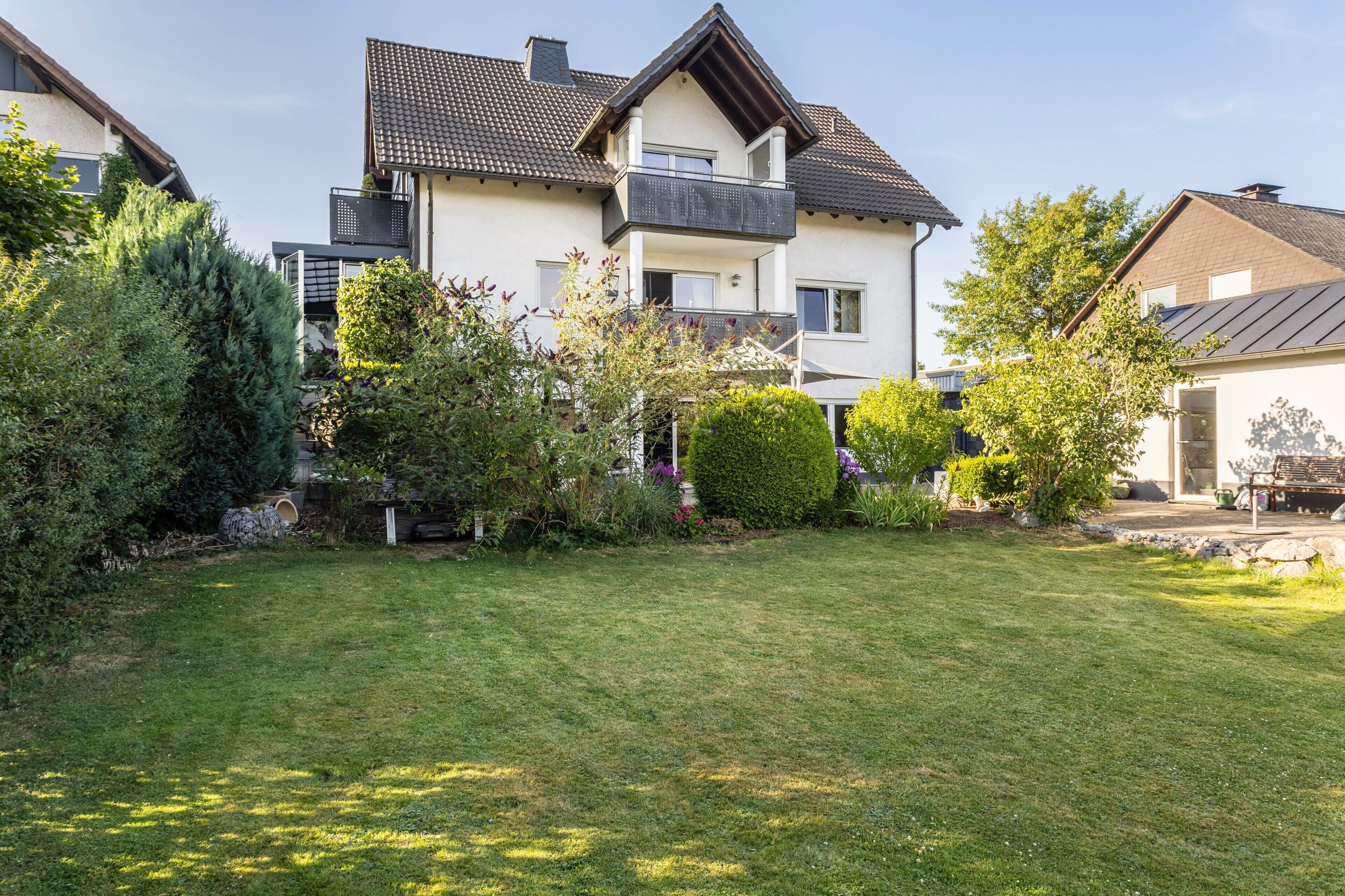 Ein zweistöckiges Wohnhaus mit Balkon, umgeben von gepflegten Gärten und einer Rasenfläche.