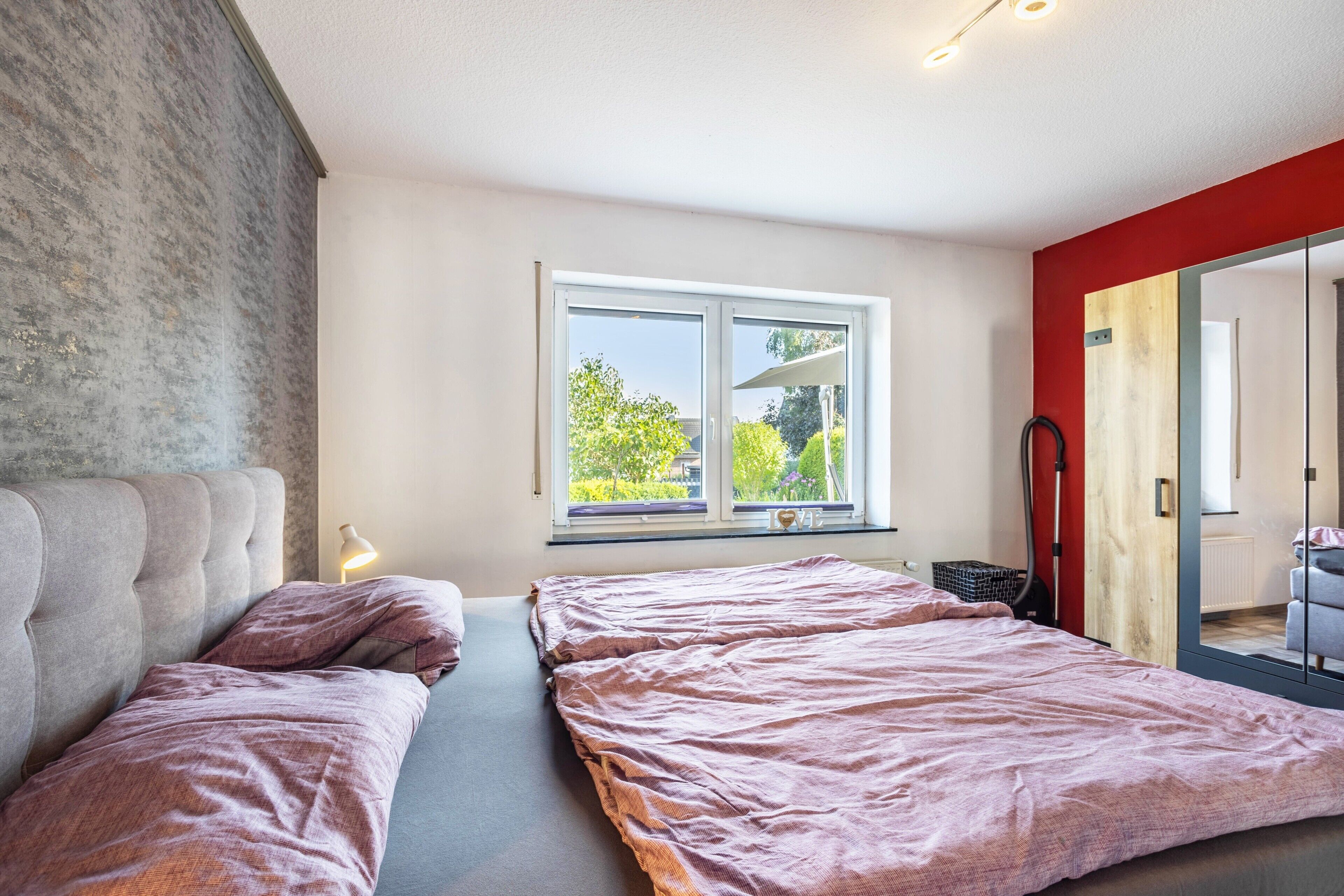 Ein Schlafzimmer mit zwei Betten, einem Fenster mit Blick auf Grünflächen und einer roten Akzentwand.
